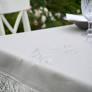 May include: A white tablecloth with a delicate lace trim and embroidered silver floral designs. The tablecloth is laid on a table with a white chair and a glass of water in the background.