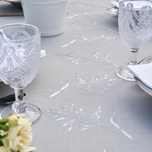 May include: A white tablecloth with a delicate floral embroidery pattern. The tablecloth is set with two glasses, a white bucket with flowers, and a white plate.