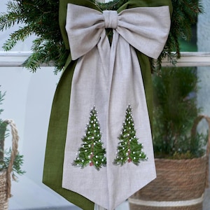 Peut inclure: Un nœud décoratif composé d'une base vert olive foncé et d'une couche supérieure beige clair. Le tissu beige présente deux sapins de Noël brodés en vert avec des accents blancs, suspendus au centre du grand nœud en tissu superposé.