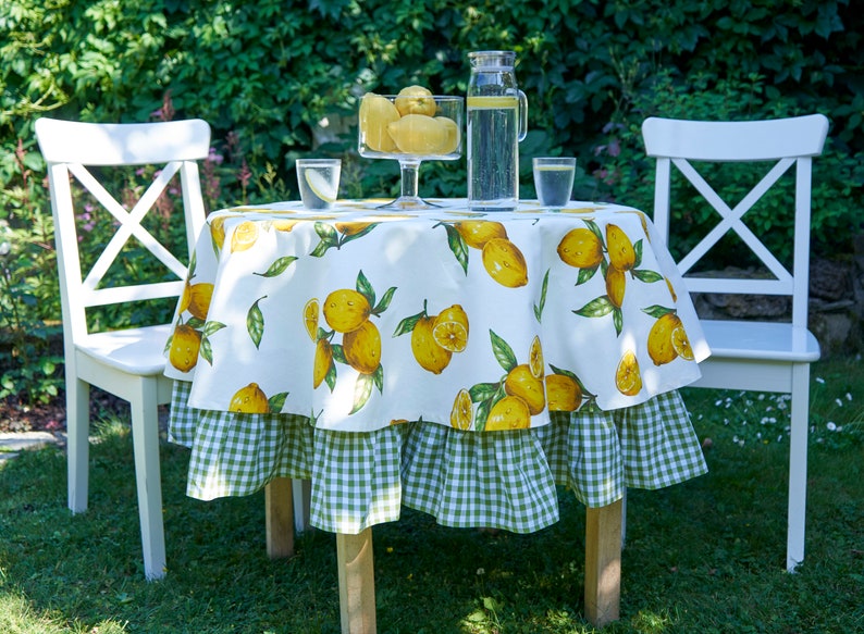 Pu&ograve; includere: Una tovaglia bianca con un bordo a quadri verdi e una stampa di limoni. La tovaglia &egrave; su un tavolo rotondo con due sedie bianche. C'&egrave; una caraffa di vetro d'acqua e una ciotola di limoni sul tavolo.