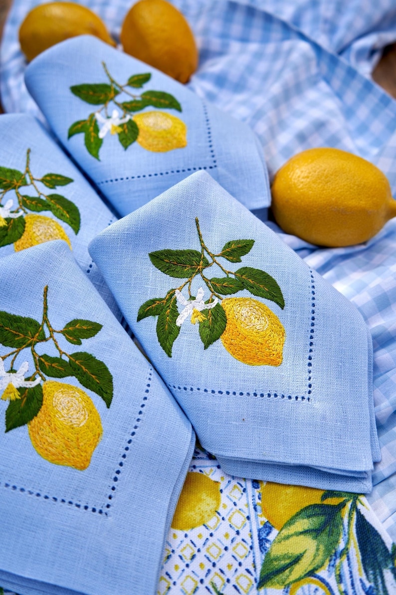 May include: Four blue linen napkins with embroidered lemon designs. The napkins are folded and stacked on top of each other. There are lemons in the background.