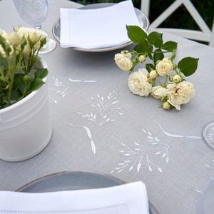 May include: A white linen tablecloth with embroidered floral designs, set with a white napkin, a gray plate, and a clear glass. A bouquet of white roses sits on the table.