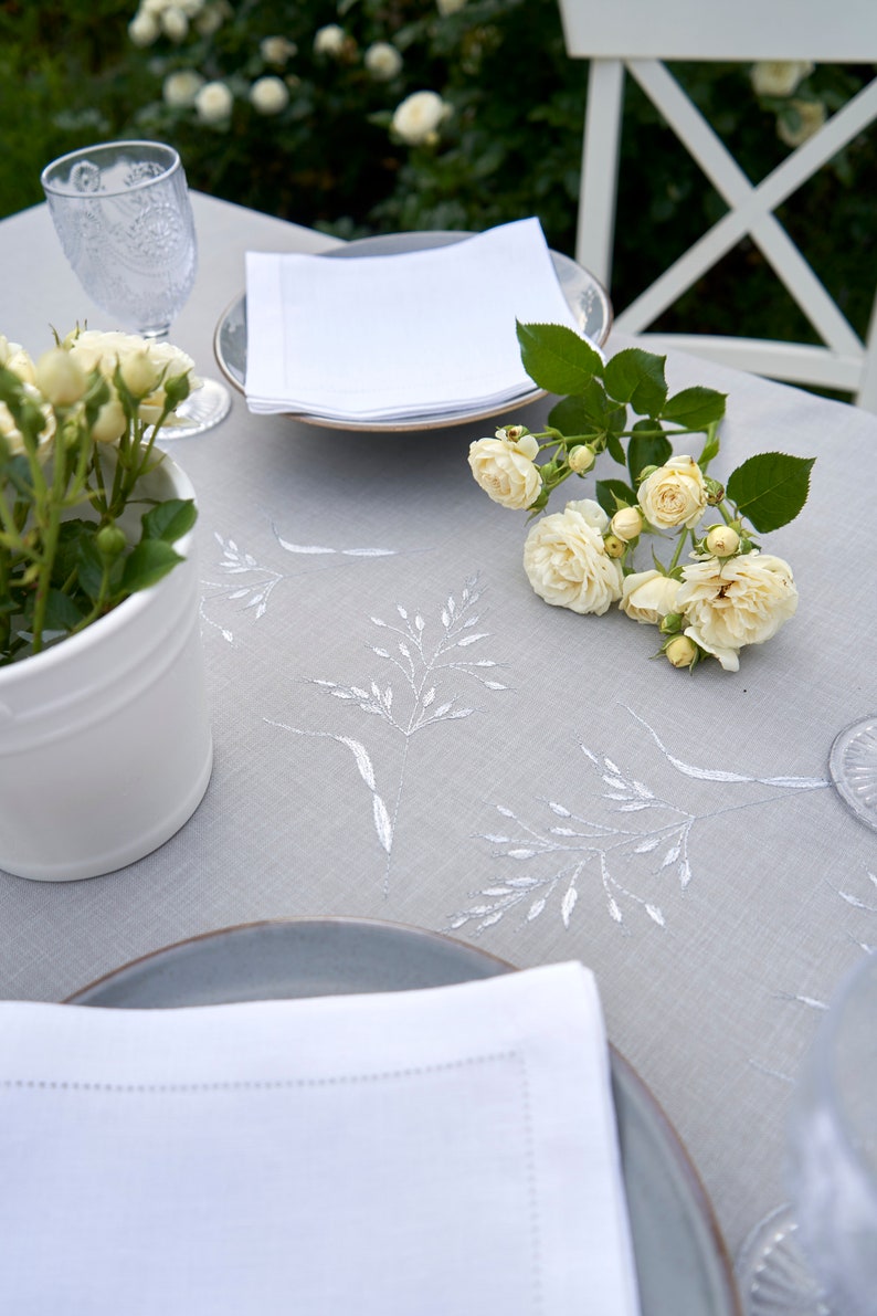 May include: A white linen tablecloth with embroidered floral designs, set with a white napkin, a gray plate, and a clear glass. A bouquet of white roses sits on the table.