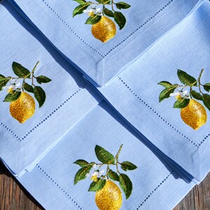 May include: Three light blue linen napkins with embroidered lemon designs. Each napkin features a yellow lemon with green leaves and white blossoms.