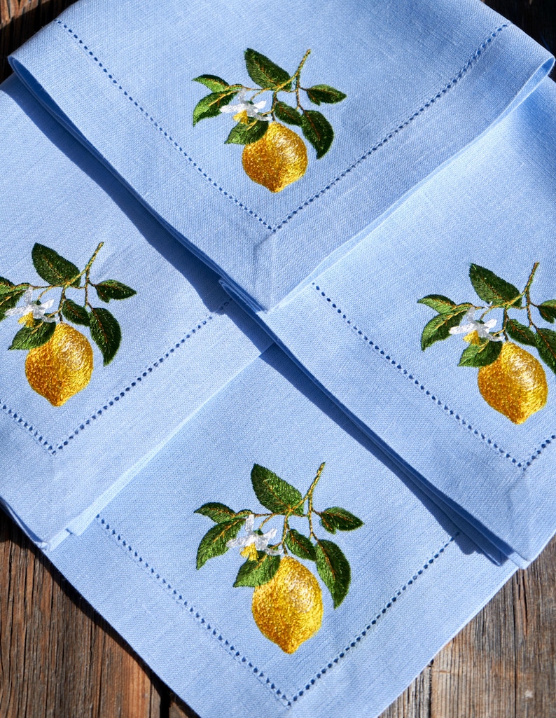 May include: Three light blue linen napkins with embroidered lemon designs. Each napkin features a yellow lemon with green leaves and white blossoms.