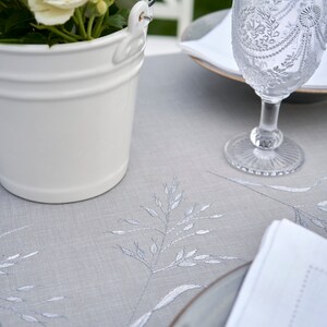 May include: A white ceramic bucket filled with white roses sits on a gray linen tablecloth with embroidered floral designs. A clear glass with a decorative pattern sits on a white napkin on a gray plate.