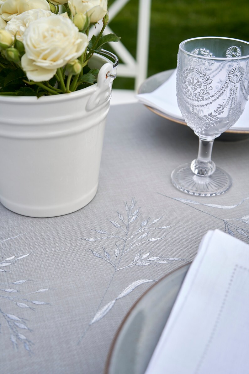 May include: A white ceramic bucket filled with white roses sits on a gray linen tablecloth with embroidered floral designs. A clear glass with a decorative pattern sits on a white napkin on a gray plate.