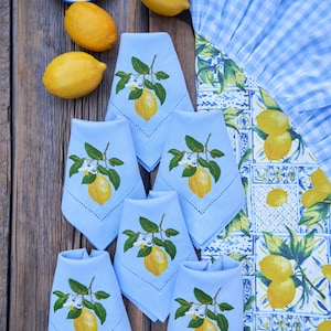 May include: Six light blue linen napkins with embroidered lemons and green leaves. The napkins are folded and arranged on a wooden table with lemons and a blue and white checkered tablecloth.