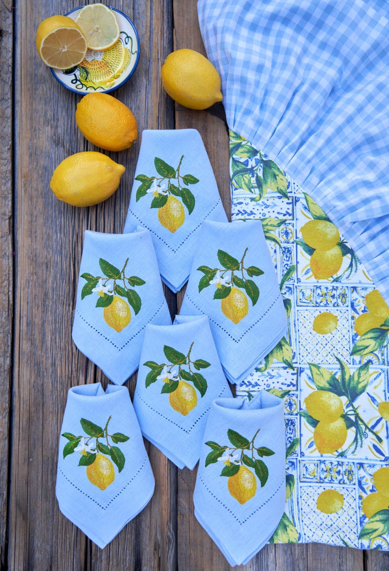 May include: Six light blue linen napkins with embroidered lemons and green leaves. The napkins are folded and arranged on a wooden table with lemons and a blue and white checkered tablecloth.