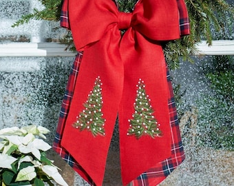 May include: A large decorative bow featuring a solid red fabric centre with two long tails, each embroidered with a green Christmas tree design. The bow is layered with a red, navy blue, and white plaid fabric trim along the edges.