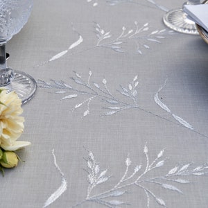 May include: A close-up of a light gray tablecloth with a delicate white embroidered floral pattern. The tablecloth is smooth and has a subtle sheen.