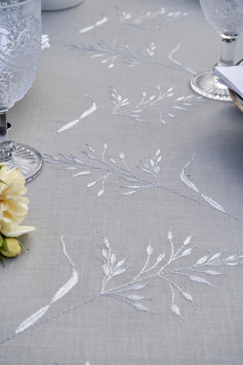 May include: A close-up of a light gray tablecloth with a delicate white embroidered floral pattern. The tablecloth is smooth and has a subtle sheen.