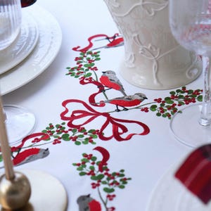 Puede incluir: Mantel blanco con bordados de cintas rojas, acebo verde y pájaros grises y rojos. Un jarrón decorativo y platos blancos están en el fondo. La mesa está decorada con un tema navideño.