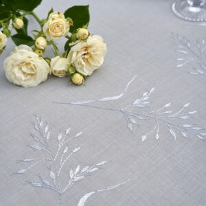 May include: A light gray linen tablecloth with a delicate embroidered design of silver-colored grass. A bouquet of white roses with green leaves is placed on the tablecloth.