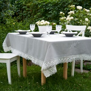 May include: A gray tablecloth with white lace trim and embroidered details is set for a dinner party in a garden setting. The table is set with white plates, clear glasses, and white chairs.