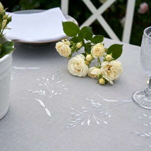 May include: A white linen tablecloth with a delicate embroidered design of grass stalks. The tablecloth is set with a white plate, a white napkin, and a clear glass. A bouquet of white roses sits on the tablecloth.
