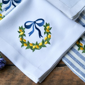 May include: White linen napkins with embroidered lemon wreaths and blue bows. The napkins are folded and arranged on a blue and white striped linen napkin.