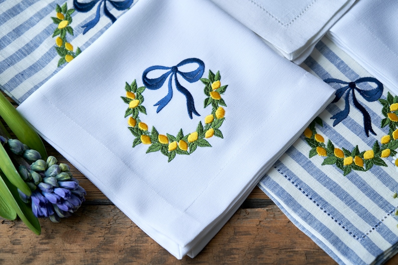 May include: White linen napkins with embroidered lemon wreaths and blue bows. The napkins are folded and arranged on a blue and white striped linen napkin.