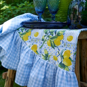 May include: A blue and white checkered table runner with a lemon print design. The runner has a ruffled edge and is displayed on a wooden table with two blue glass goblets and a blue plate.