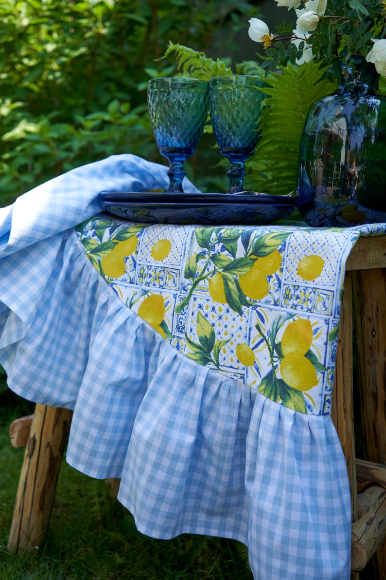 May include: A blue and white checkered table runner with a lemon print design. The runner has a ruffled edge and is displayed on a wooden table with two blue glass goblets and a blue plate.