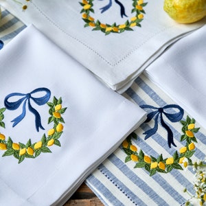 May include: Four white linen napkins with blue bows and embroidered lemon wreaths. The napkins are folded and arranged on a blue and white striped tablecloth.