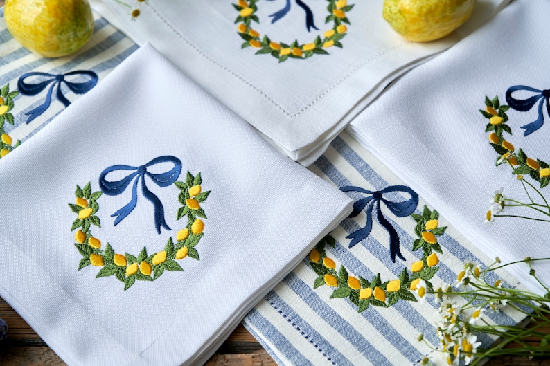 May include: Four white linen napkins with blue bows and embroidered lemon wreaths. The napkins are folded and arranged on a blue and white striped tablecloth.