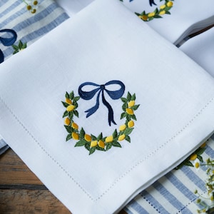 May include: White linen napkins with embroidered lemon wreaths and blue bows. The napkins are folded and arranged on a blue and white striped surface.