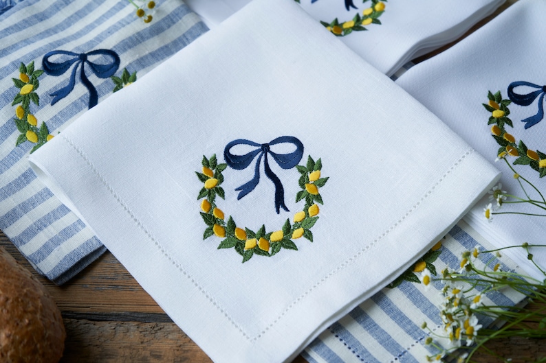 May include: White linen napkins with embroidered lemon wreaths and blue bows. The napkins are folded and arranged on a blue and white striped surface.