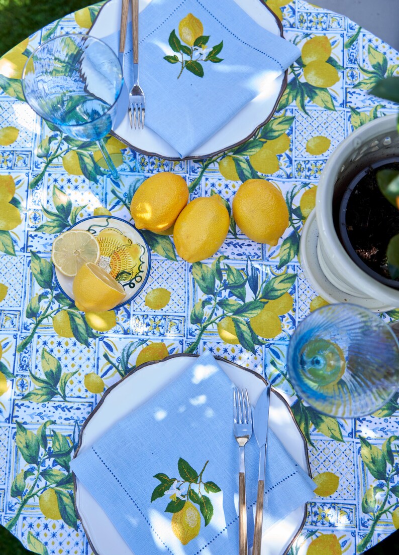 May include: A blue and white tablecloth with a lemon pattern. There are two place settings with blue napkins, white plates, and wooden utensils. There are lemons on the table and a potted lemon tree in the background.