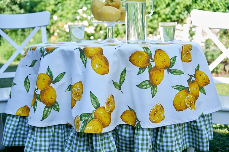 Pu&ograve; includere: Tovaglia rotonda bianca con un bordo a quadri verde e bianco. La tovaglia presenta una stampa di limone con limoni gialli e foglie verdi.