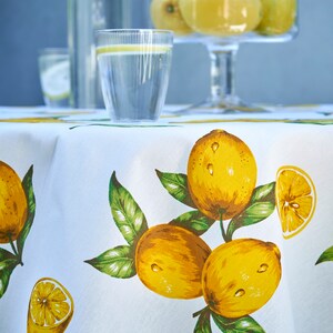 Pu&ograve; includere: Tovaglia bianca con stampa di limone. La tovaglia presenta limoni gialli con foglie verdi. Ci sono due bicchieri d'acqua e una caraffa d'acqua sul tavolo.