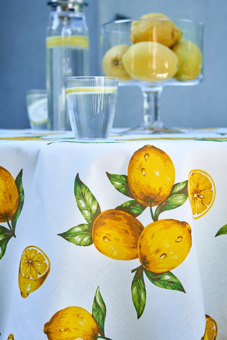 Pu&ograve; includere: Tovaglia bianca con stampa di limone. La tovaglia presenta limoni gialli con foglie verdi. Ci sono due bicchieri d'acqua e una caraffa d'acqua sul tavolo.