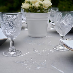 May include: A white tablecloth with an embroidered floral pattern.  Two clear glass wine glasses with intricate etched designs are on the table, along with a white ceramic planter with white flowers.