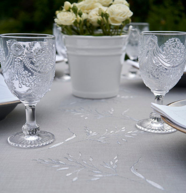 May include: A white tablecloth with an embroidered floral pattern.  Two clear glass wine glasses with intricate etched designs are on the table, along with a white ceramic planter with white flowers.