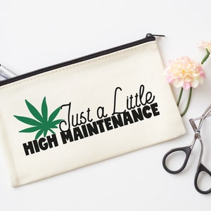 Puede incluir: Bolsa de maquillaje de lona color crema con cremallera negra y el texto negro "Just a Little High Maintenance". Un gráfico de hoja de cannabis verde a la izquierda. Pinceles de maquillaje y un rizador de pestañas.