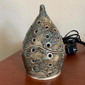 May include: A brown and green ceramic electric oil diffuser with a pointed top and a speckled glaze. The diffuser has a cord and plug.