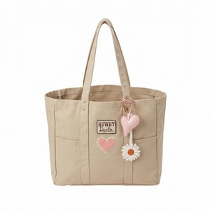 May include: A beige canvas tote bag with two exterior pockets. The bag features patches that read "HOWDY DARLIN" and a pink heart. A pink heart and white daisy charm hang from the handle. The bag has two handles for carrying.