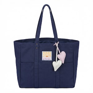 May include: A navy blue tote bag with two exterior pockets and a rectangular patch with the text "TANNER & TIPSY". The bag has two handles and a charm with two heart-shaped pendants, one pink and one white.