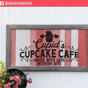Op de afbeelding: Een houten bord met een rood-wit-grijze gestreepte achtergrond. Het bord luidt "Cupid's Cupcake Cafe - Baked with love in every bite".