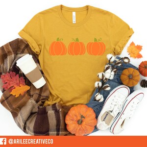 Peut inclure: Un t-shirt jaune moutarde avec un motif de trois citrouilles orange avec des tiges vertes. Le t-shirt est pos&eacute; sur une couverture en plaid brun et blanc avec une tasse &agrave; caf&eacute; blanche, des feuilles d'automne et une paire de baskets blanches.