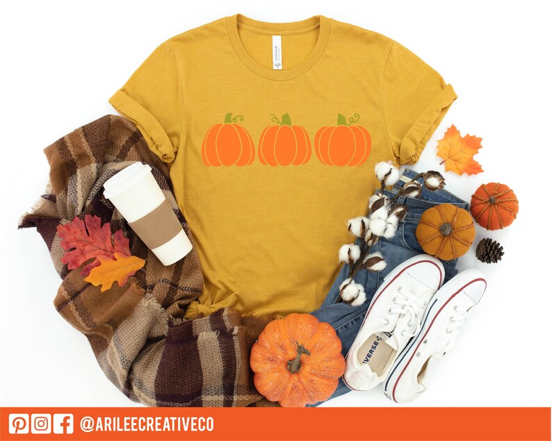 Peut inclure: Un t-shirt jaune moutarde avec un motif de trois citrouilles orange avec des tiges vertes. Le t-shirt est pos&eacute; sur une couverture en plaid brun et blanc avec une tasse &agrave; caf&eacute; blanche, des feuilles d'automne et une paire de baskets blanches.
