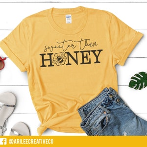 Peut inclure: Un t-shirt jaune avec le texte "Sweeter Than Honey" et un graphique d'abeille. Le t-shirt est sur une surface en bois blanc avec une paire de sandales, des lunettes de soleil et un porte-cl&eacute;s en forme de feuille verte.