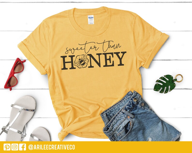 Peut inclure: Un t-shirt jaune avec le texte "Sweeter Than Honey" et un graphique d'abeille. Le t-shirt est sur une surface en bois blanc avec une paire de sandales, des lunettes de soleil et un porte-cl&eacute;s en forme de feuille verte.