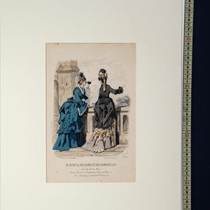 Puede incluir: Una impresi&oacute;n antigua titulada "LE JOURNAL DES DAMES ET DES DEMOISELLES" representa a dos mujeres con vestidos de &eacute;poca. Una lleva un vestido azul y la otra un vestido negro con un detalle amarillo. La impresi&oacute;n est&aacute; enmarcada en blanco.
