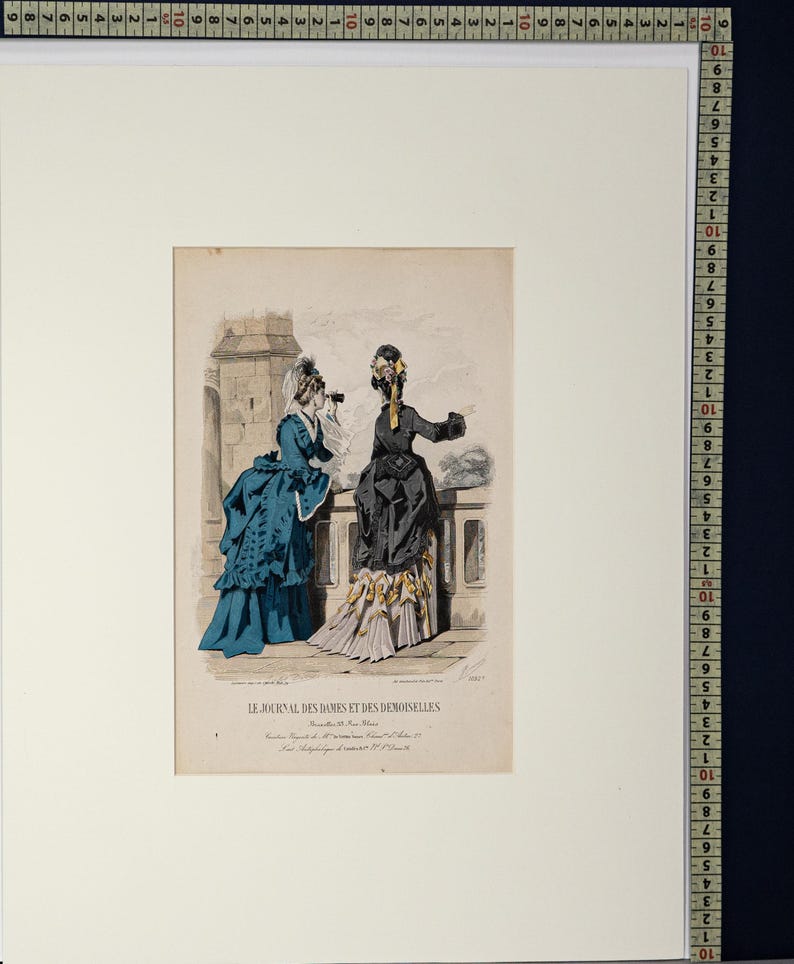 Puede incluir: Una impresi&oacute;n antigua titulada "LE JOURNAL DES DAMES ET DES DEMOISELLES" representa a dos mujeres con vestidos de &eacute;poca. Una lleva un vestido azul y la otra un vestido negro con un detalle amarillo. La impresi&oacute;n est&aacute; enmarcada en blanco.