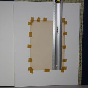 Puede incluir: Un trozo rectangular de papel de color beige est&aacute; pegado a una superficie blanca con peque&ntilde;os trozos de cinta adhesiva amarilla. Una regla plateada con marcas negras y un nivel se coloca verticalmente sobre el papel. El papel est&aacute; rodeado de hojas blancas.