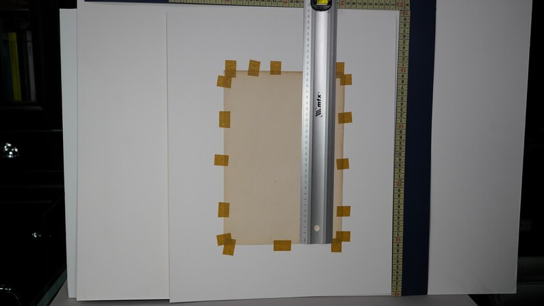 Puede incluir: Un trozo rectangular de papel de color beige est&aacute; pegado a una superficie blanca con peque&ntilde;os trozos de cinta adhesiva amarilla. Una regla plateada con marcas negras y un nivel se coloca verticalmente sobre el papel. El papel est&aacute; rodeado de hojas blancas.