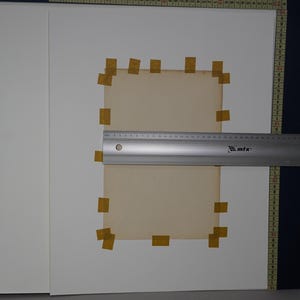 Puede incluir: Una regla de metal plateado con un nivel est&aacute; midiendo un papel rectangular de color marr&oacute;n claro pegado a una superficie blanca. El papel est&aacute; rodeado de trozos de cinta adhesiva beige. El fondo incluye papel blanco y una gu&iacute;a de medici&oacute;n azul oscuro.