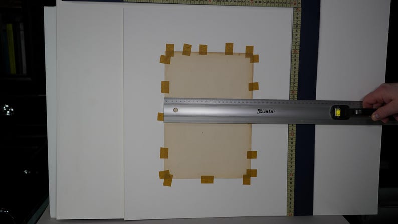 Puede incluir: Una regla de metal plateado con un nivel est&aacute; midiendo un papel rectangular de color marr&oacute;n claro pegado a una superficie blanca. El papel est&aacute; rodeado de trozos de cinta adhesiva beige. El fondo incluye papel blanco y una gu&iacute;a de medici&oacute;n azul oscuro.