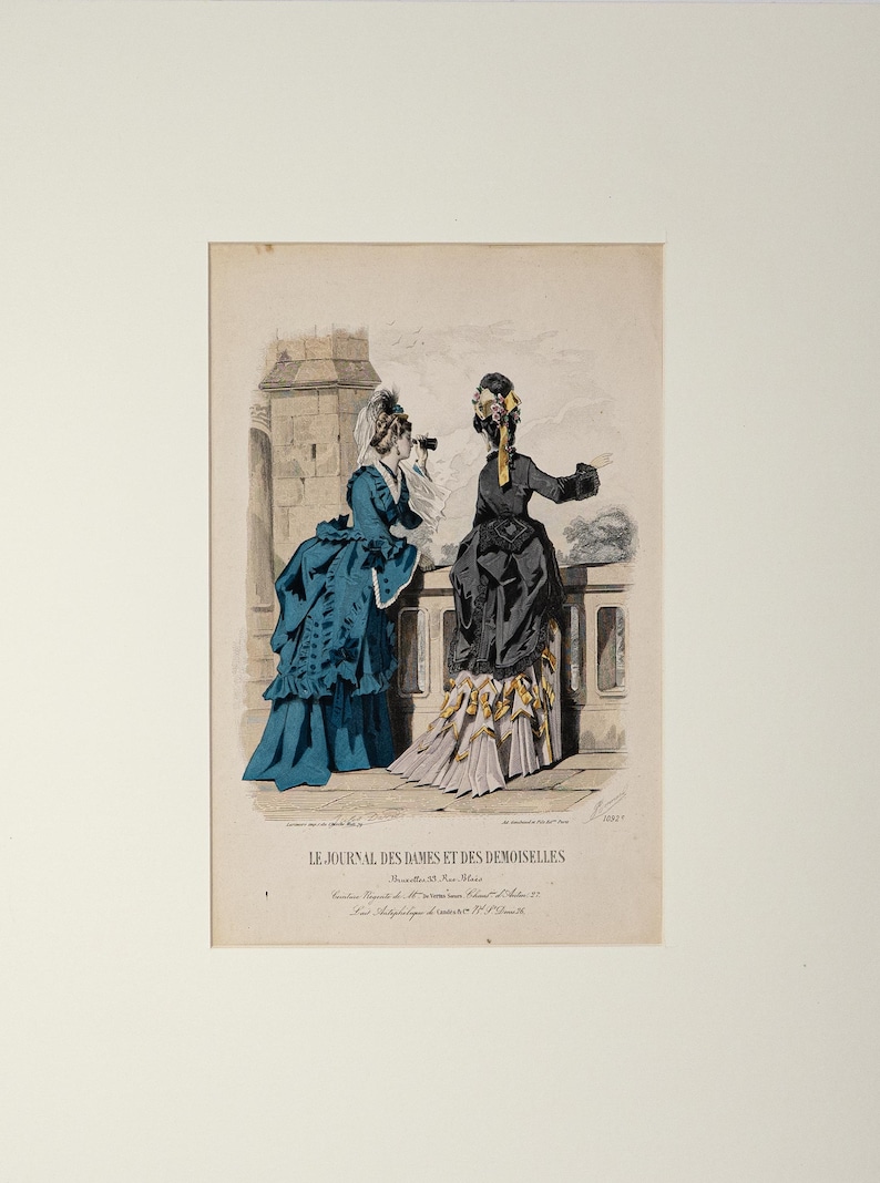 Puede incluir: Una impresi&oacute;n antigua titulada "Le Journal des Dames et des Demoiselles" con dos mujeres con vestidos elaborados en un balc&oacute;n. Una mujer lleva un vestido verde azulado, la otra un vestido negro con detalles amarillos. La impresi&oacute;n est&aacute; enmarcada.